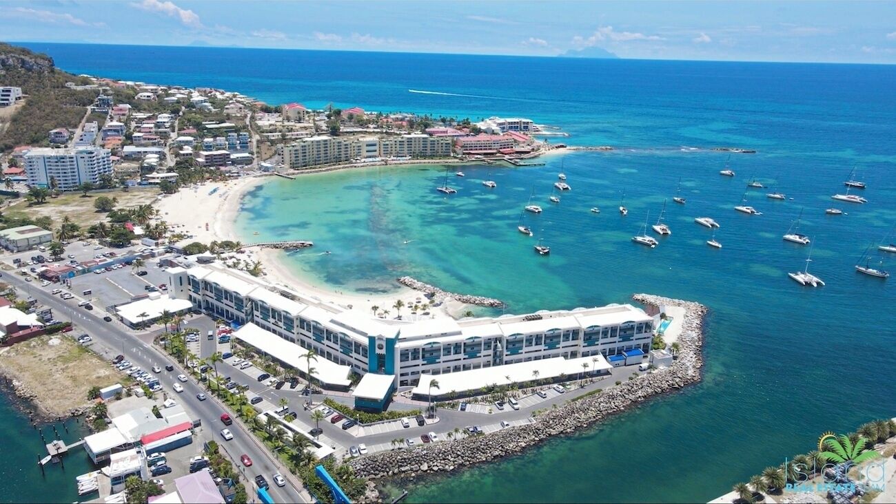 An aerial view of a coastal resort with a beachfront hotel, clear blue waters, and numerous boats anchored nearby.