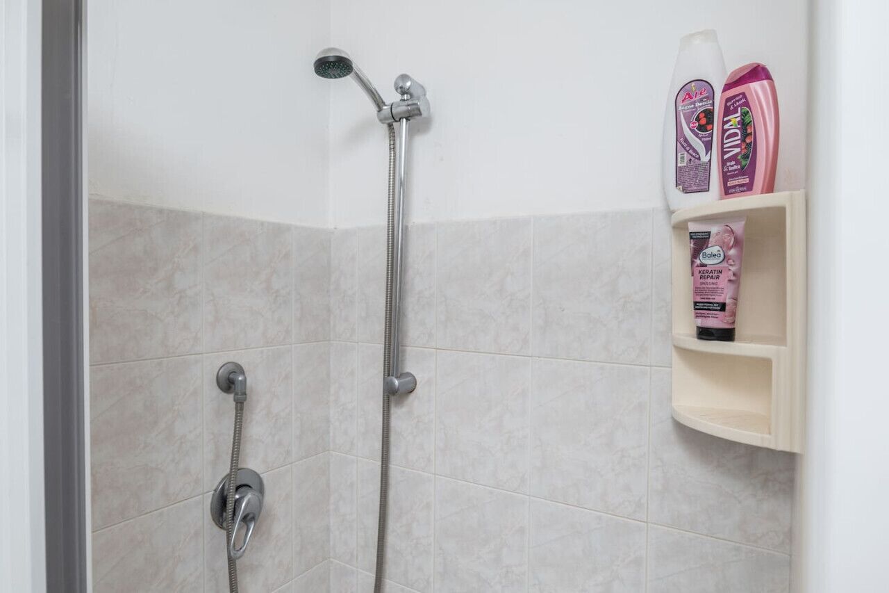 Un espace douche équipé d’un pommeau de douche à main et d’un robinet mural. On y trouve des articles de toilette sur une étagère, notamment un flacon de shampoing et un flacon d’après-shampoing.