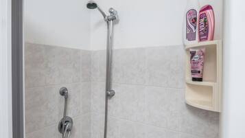Un espace douche équipé d’un pommeau de douche à main et d’un robinet mural. On y trouve des articles de toilette sur une étagère, notamment un flacon de shampoing et un flacon d’après-shampoing.