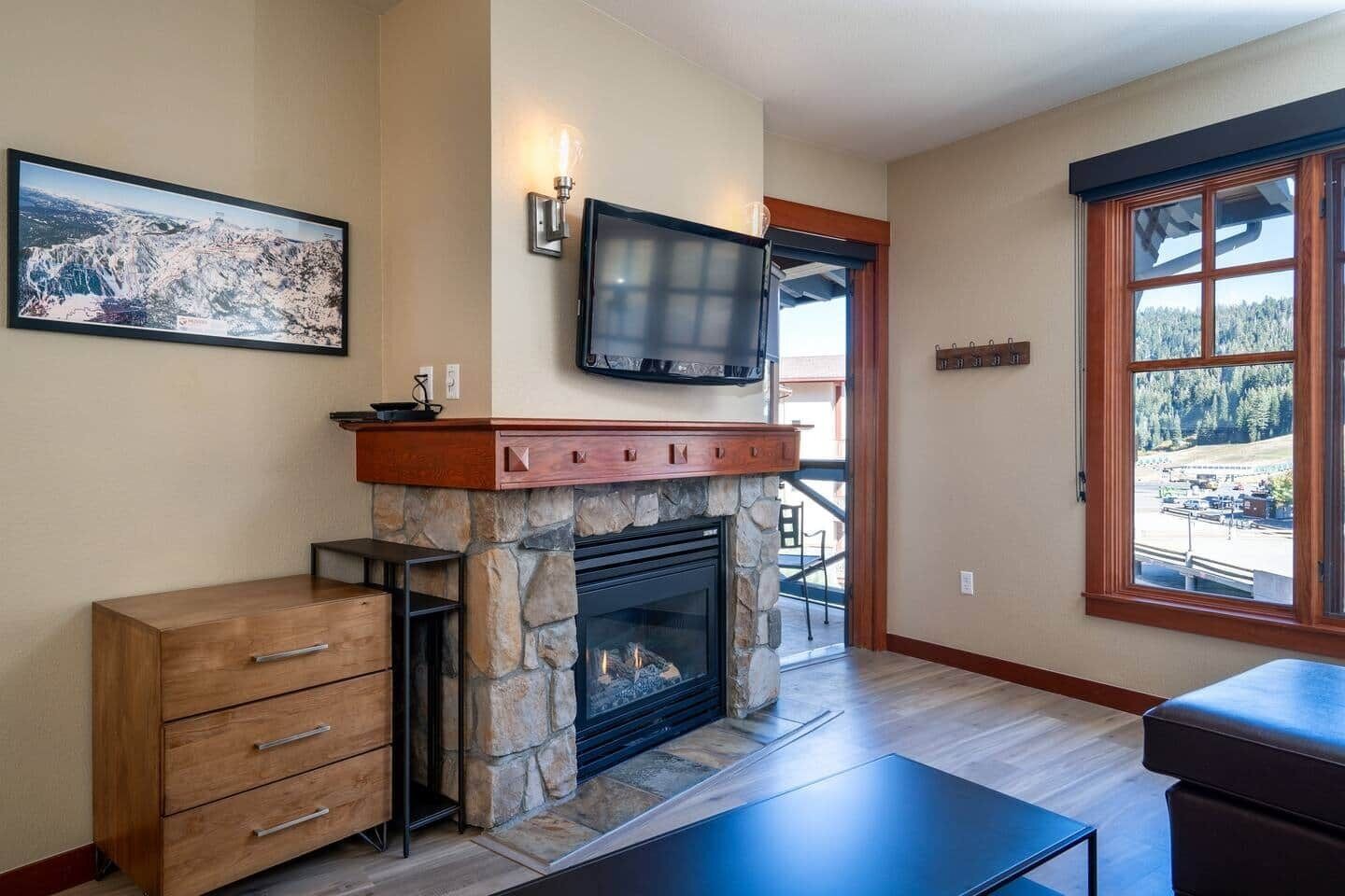A living room with a stone fireplace, a television mounted above it, and a large window offering a view of a forested area.