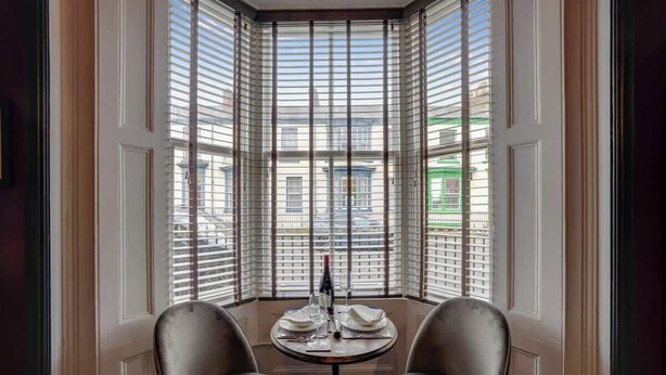 Une chambre d’angle avec une table dressée pour deux personnes, de grandes fenêtres équipées de stores et une vue sur les bâtiments environnants.