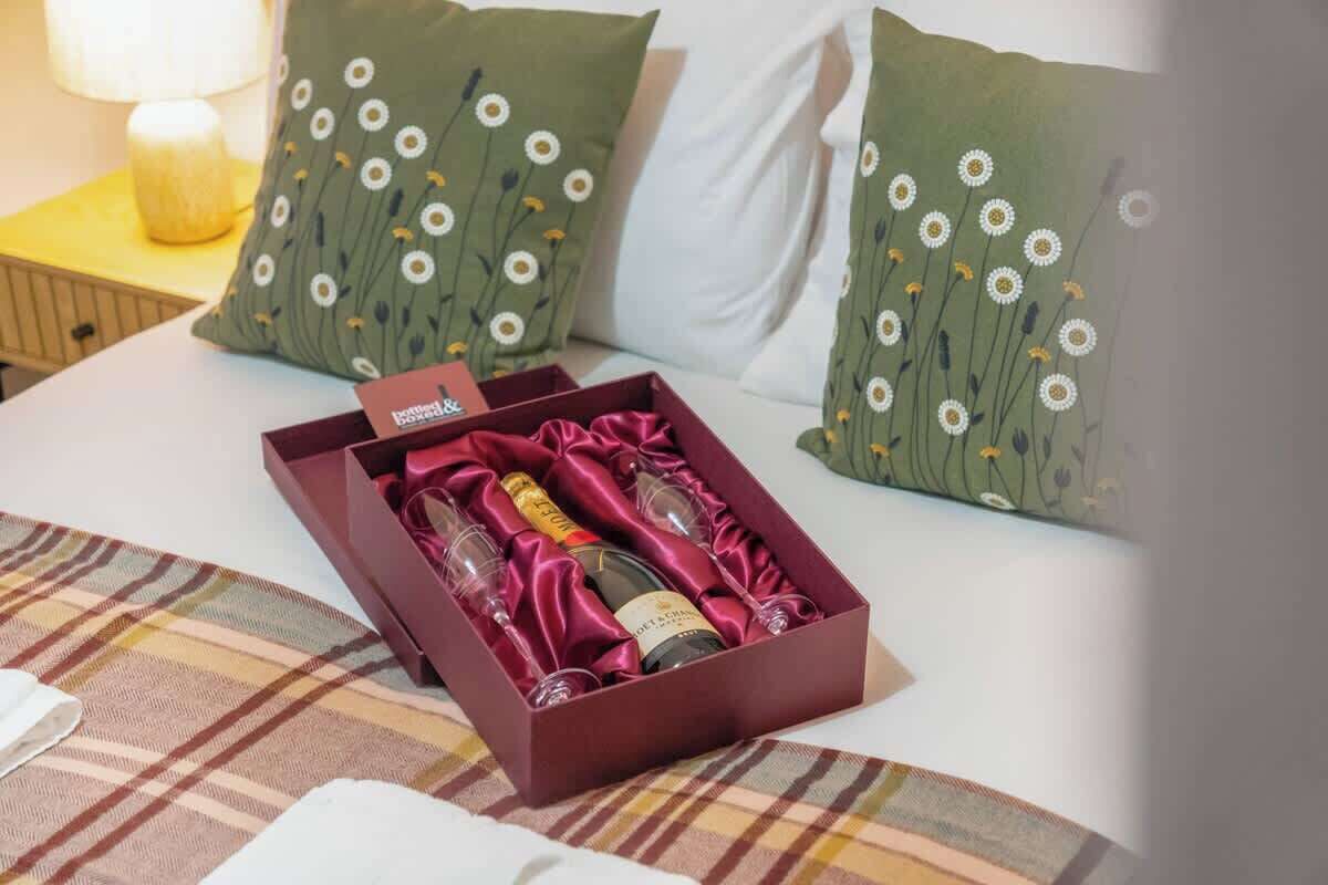 A bed with a plaid blanket, a box of wine, and two pillows with a floral pattern.