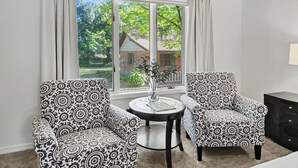 A room with two patterned armchairs, a small round table with a vase, and a view of a house through a large window. - Spacious & Lovely 4-BR Home Minutes to WEC, HITS (Ocala)