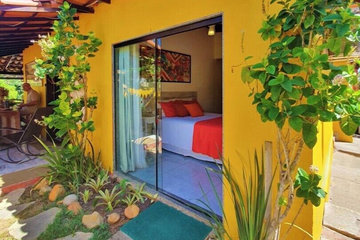 Edifício amarelo com porta de vidro que dá para um quarto com cama e manta vermelha. Há plantas e um pequeno jardim com pedras e tapete verde.
