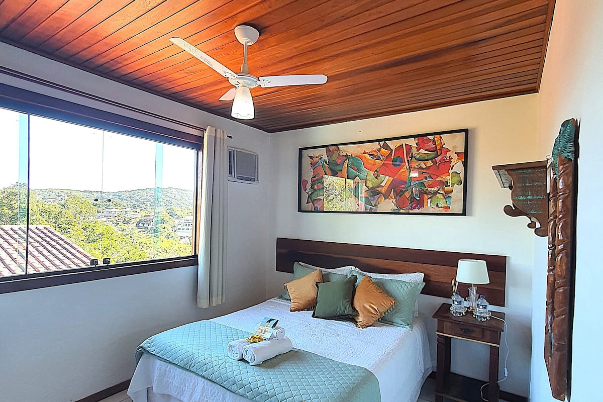 Quarto com cama, mesa de cabeceira, ventoinha no teto, janela com vista e um quadro na parede.