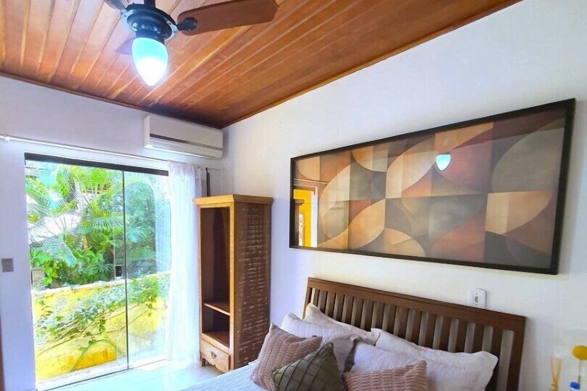 Quarto com cama, ventoinha no teto, janela grande com vista para vegetação e pintura abstrata pendurada na parede.