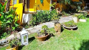 Un bâtiment jaune avec un toit recouvert de tuiles, entouré d’un jardin bien entretenu où poussent différentes espèces de plantes et un palmier.