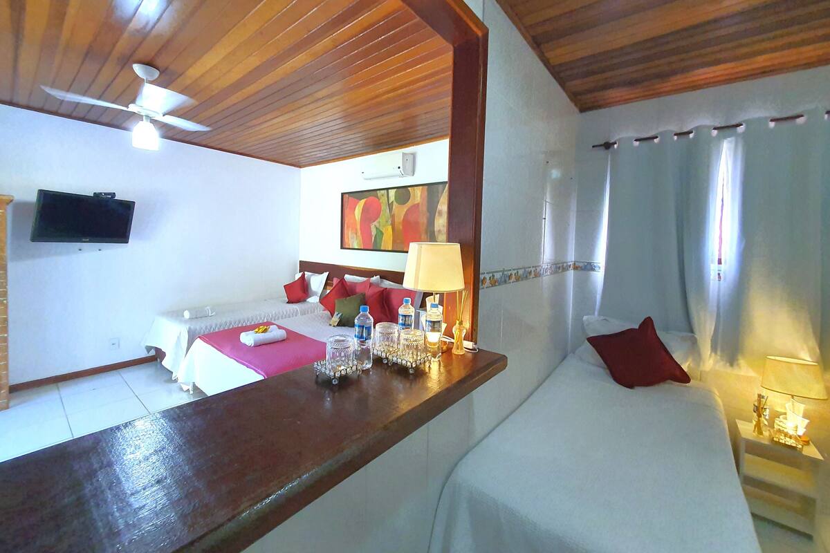 Quarto de hotel com teto de madeira, cama com roupa de cama branca, mesa de cabeceira com abajur, televisão na parede e um quadro sobre a cama.