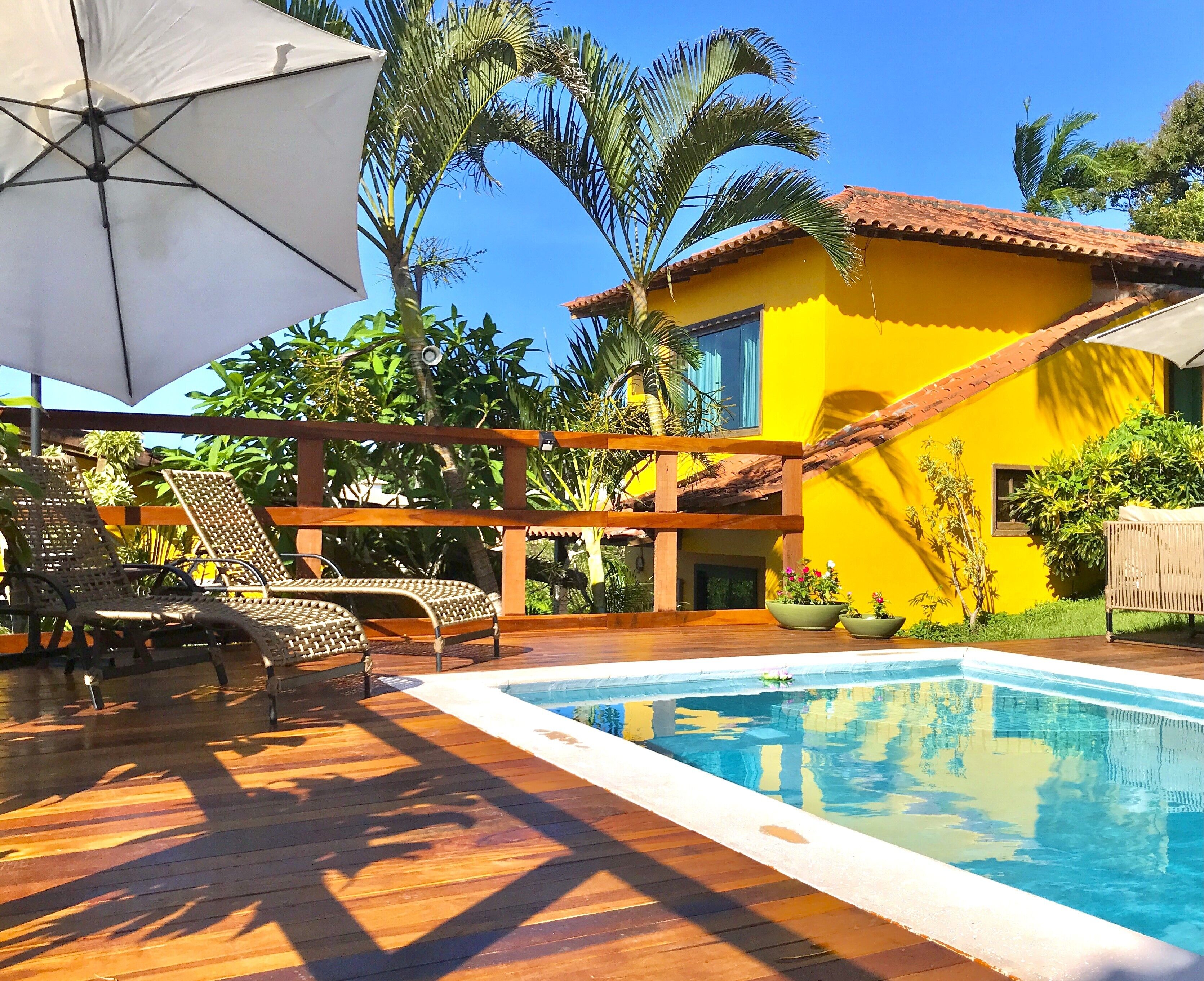 A pool area with lounge chairs and an umbrella, surrounded by lush greenery and a yellow building.