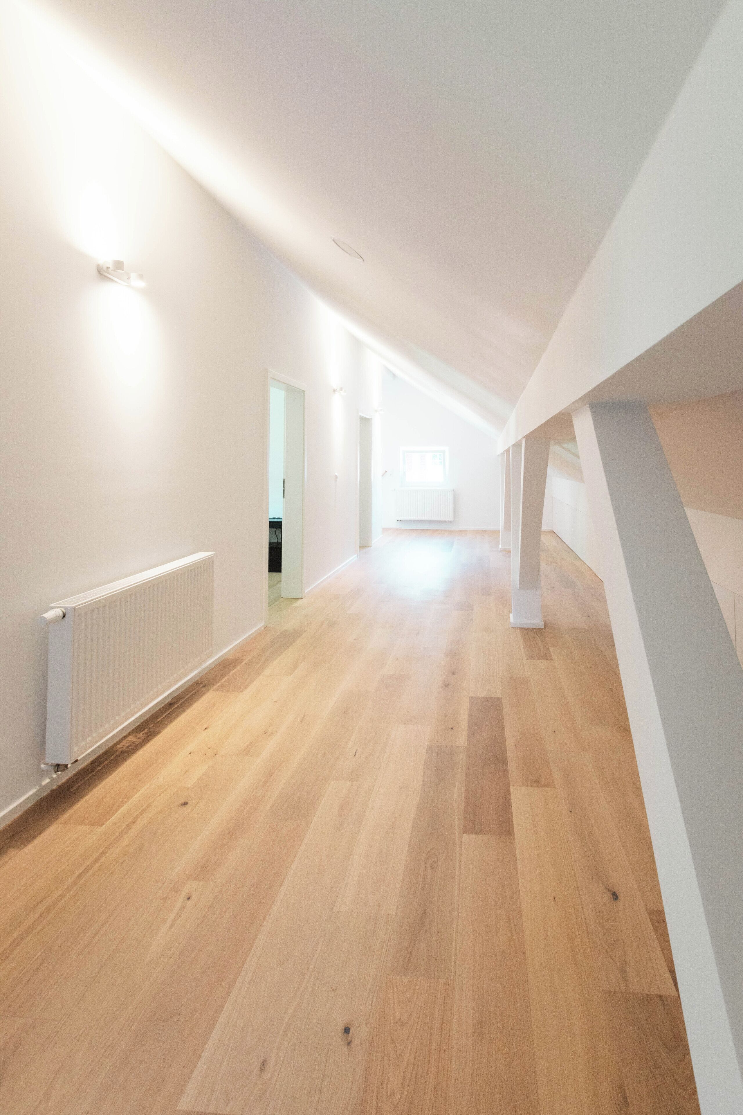 A spacious, well-lit corridor with wooden flooring and white walls, leading to a room with a window and a radiator.
