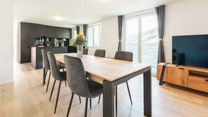 A modern kitchen with dark cabinets, a wooden dining table with chairs, a TV on a wooden stand, and a balcony with a view of the mountains.