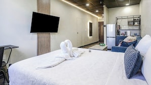Habitación de hotel con cama, televisor de pantalla plana en la pared, kitchenette con refrigerador y una pequeña mesa.