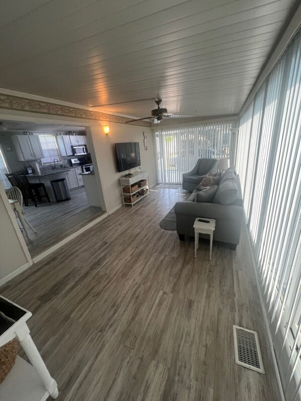 A spacious living area with a grey sofa, a small white side table, and a television. A kitchen is visible in the background with white cabinets and a microwave. The room has a wooden floor and large windows with blinds. - Enjoy a tropical vacation at this destination rental. (Jensen Beach)