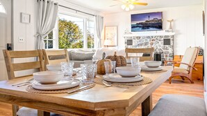 A dining area with a wooden table set for four, chairs, a stone fireplace, and a television above it. - Salt Box by the Sea: Water View - Charming 3-bedroom Cottage in Wells! (Wells)