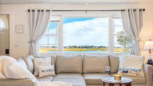 A cozy living room with a large window, a beige sofa, and a small table with glasses and a candle. - Salt Box by the Sea: Water View - Charming 3-bedroom Cottage in Wells! (Wells)