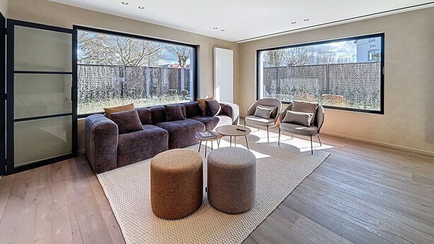 A spacious living room with a large sectional sofa, a coffee table, and two armchairs. The room features large windows offering a view of the outdoors.