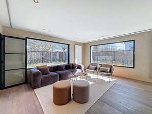 Een ruime woonkamer met een grote hoekbank, een salontafel en twee fauteuils. De kamer heeft grote ramen met uitzicht op de omgeving.