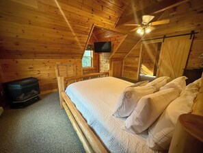 A wooden cabin bedroom with a bed, a ceiling fan, a television, and a fireplace. - Fernwood | Secluded | Deep Creek & Wisp | Hot Tub (Accident)