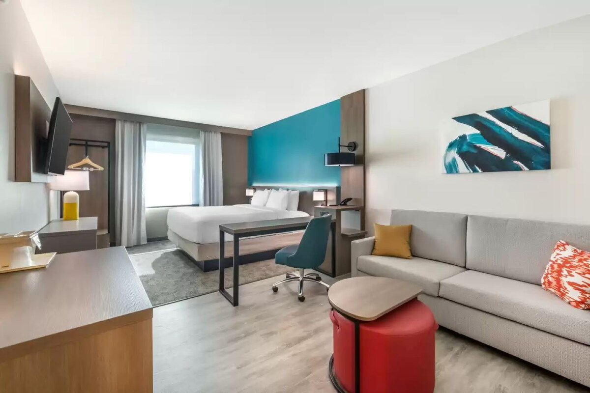 A modern hotel room with a bed, desk, chair, and a red suitcase.