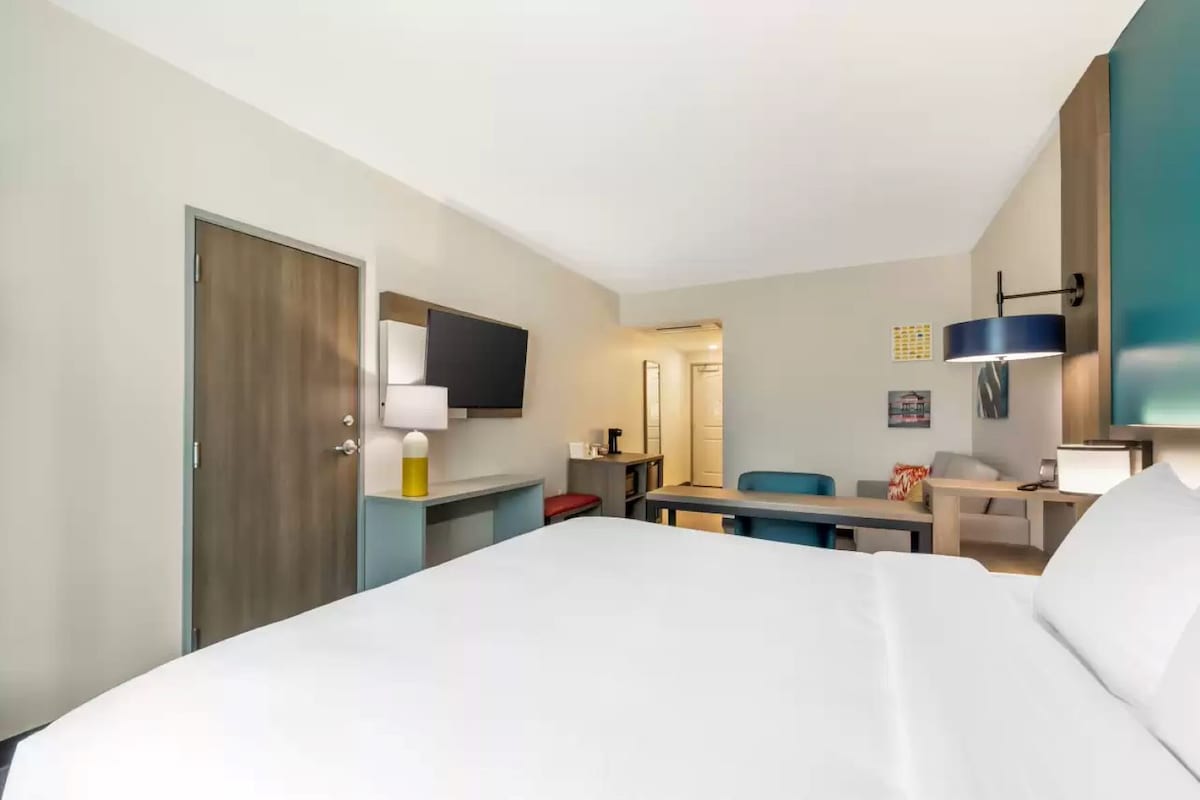A hotel room with a large bed, a desk, a chair, a TV, and a lamp.