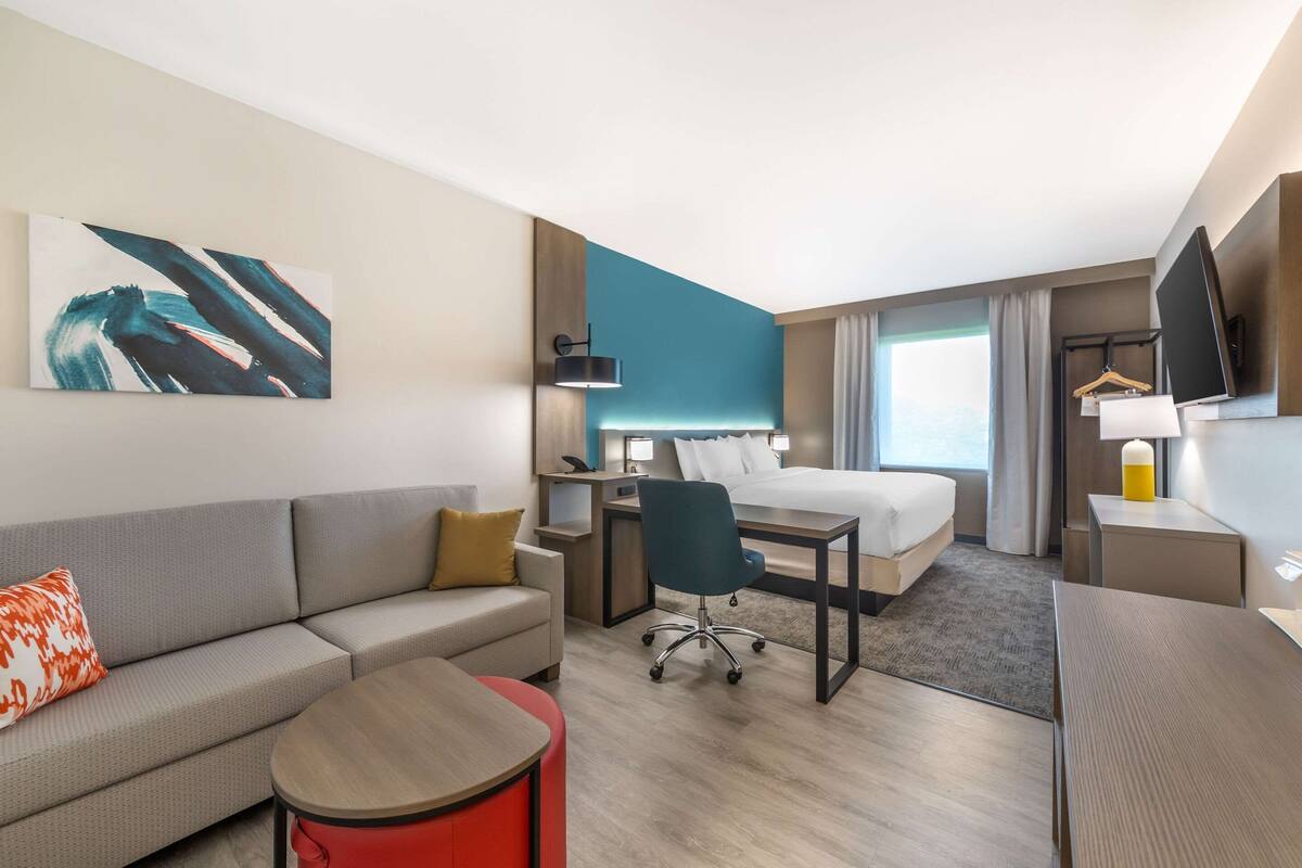 A modern hotel room with a bed, a desk, a sofa, a chair, and a TV.