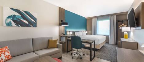 A modern hotel room with a bed, a desk, a sofa, a chair, and a TV.