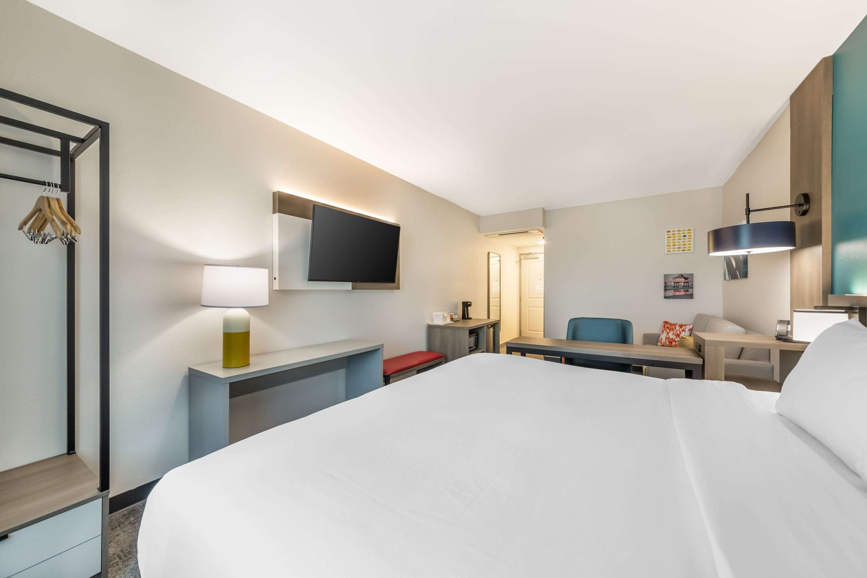 A hotel room with a large bed, a desk with a chair, a TV mounted on the wall, and a bedside table with a lamp.