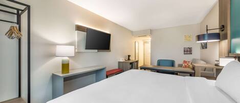 Una habitación de hotel con una cama grande, un escritorio con silla, un televisor montado en la pared y una mesita de noche con lámpara.