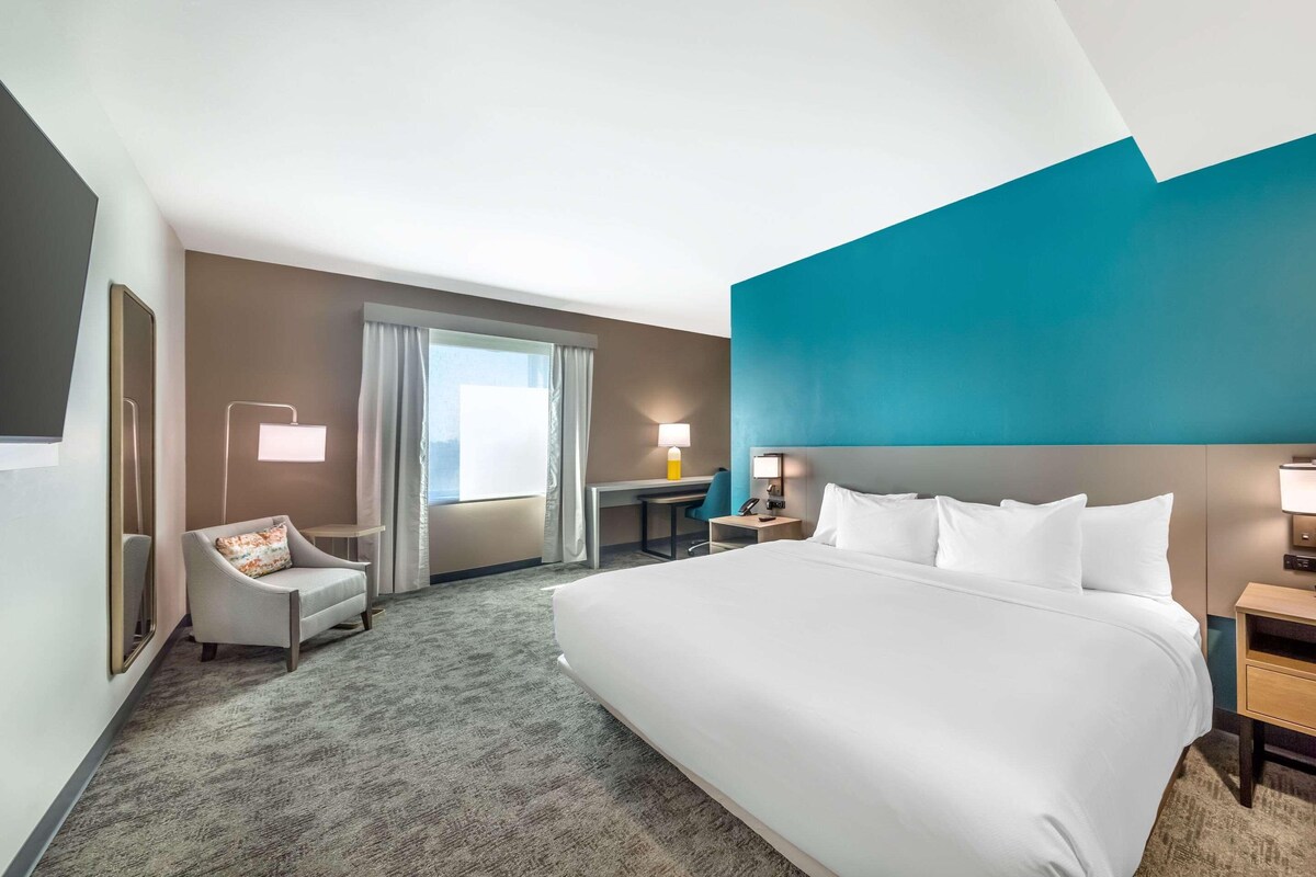 Habitación de hotel con una cama grande, un escritorio, una silla y vistas al exterior a través del ventanal.