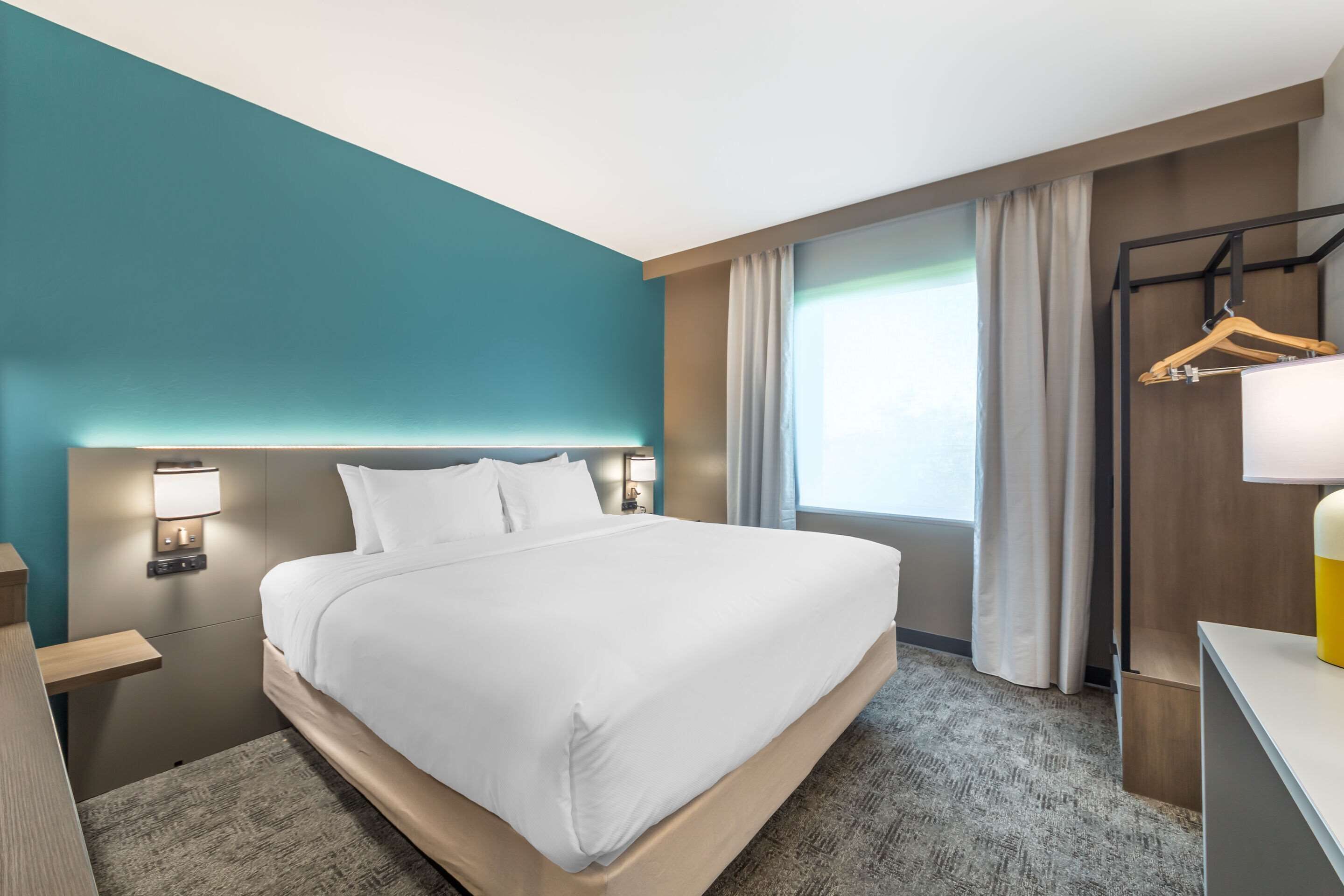 Habitación de hotel con una cama grande, una mesita de noche con lámpara, un aparador de madera, un escritorio con lámpara y vistas al exterior a través de una ventana.