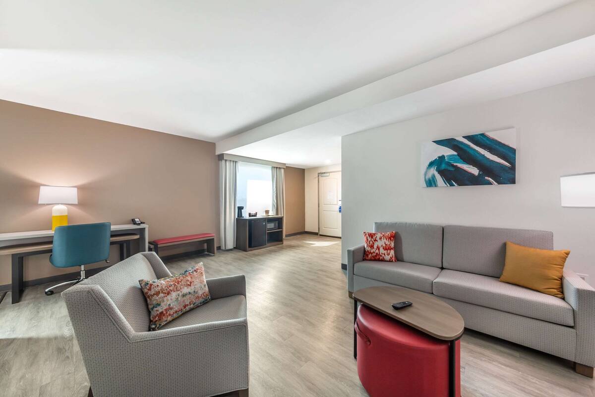 A modern hotel room with a grey sofa, a red ottoman, a desk with a lamp, and a large abstract painting on the wall.