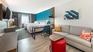 A modern hotel room with a bed, a desk, a sofa, and a red ottoman. - Comfort Suites Fort Myers East I-75 (Fort Myers)