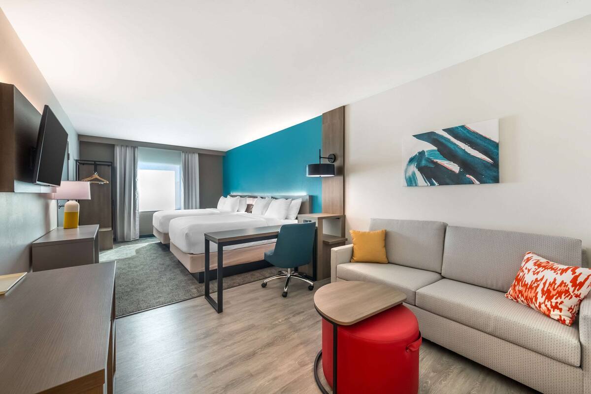 A modern hotel room with a bed, a desk, a sofa, and a red ottoman.