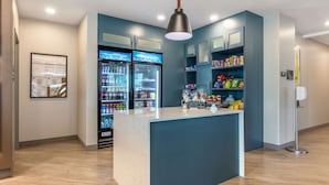 A modern hotel lobby with a central counter, two refrigerators stocked with beverages, and shelves with snacks. - Comfort Suites Fort Myers East I-75 (Fort Myers)