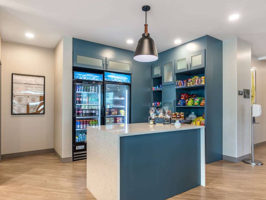 Un moderno vestíbulo de hotel con un mostrador central, dos refrigeradores llenos de bebidas y estantes con aperitivos.