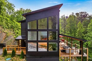 A modern cabin with large windows, a deck, and a view of the surrounding forest. - New! Mt.View! HotTub! Close to Dollywood! FirePit! (Sevierville)