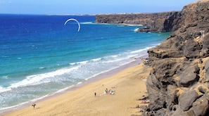 A beach with a rocky cliff, a person with a kite, and a few people on the sand.