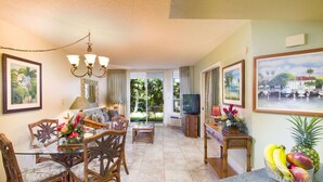 A well-lit living room with a television, dining area with chairs and a table, and artwork on the walls. - Maui Banyan - 1BR Partial Ocean View - No Cleaning Fee/ Full Kitchen (Kihei)