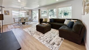 A modern living room with a sectional sofa, a dining area with a table and chairs, and large windows offering a view of the outdoors. - Airy Modern Farmhouse 5BR/3BA Oasis w/ Fast Wi-Fi (Kent)