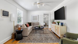 A modern living room with a sectional sofa, a coffee table, a TV mounted on the wall, and a cabinet with decorative items. - Park Circle - Firefly - Renovated 3 Bedroom! (North Charleston)