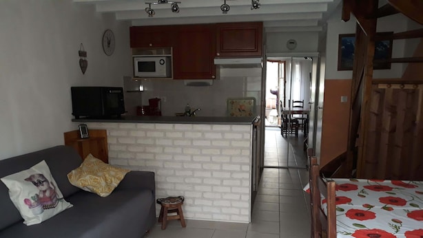 A compact living space with a sofa, a small stool, a dining table with a floral tablecloth, and a kitchen area with a microwave and cabinets. - 2* apartment in the mountains (Luz-Saint-Sauveur)