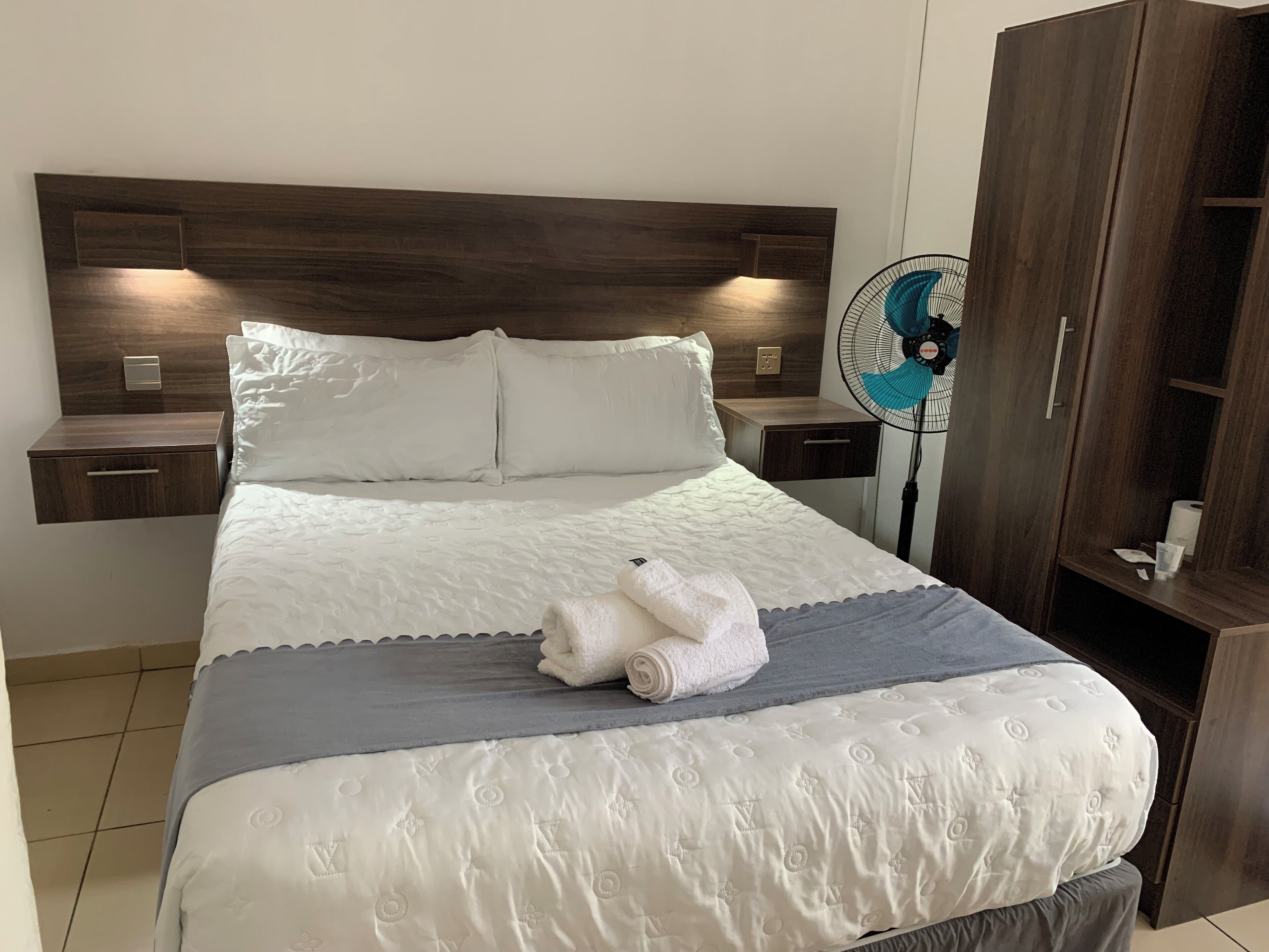 Un lit bien fait, avec une couette blanche, un plaid gris et une serviette blanche enroulée. On y voit une tête de lit en bois, une table de chevet avec un tiroir et un ventilateur sur pied.