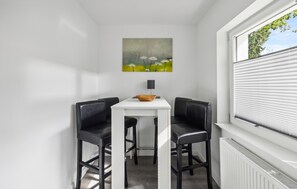 A small dining area with a white table and two black chairs, a painting of flowers on the wall, and a window with blinds.