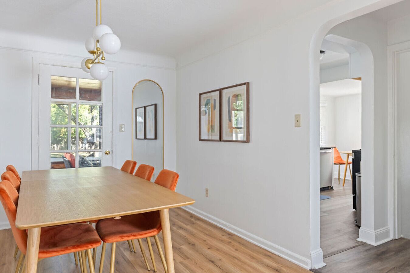 A dining area with a wooden table and orange chairs, a white wall with framed pictures, and a view through a window.