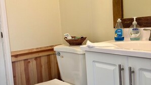 A bathroom with a toilet, a wooden panel wall, a white sink with a mirror, and toiletries. - Waterfront Bunkroom sits on the Colorado river. (Matagorda)