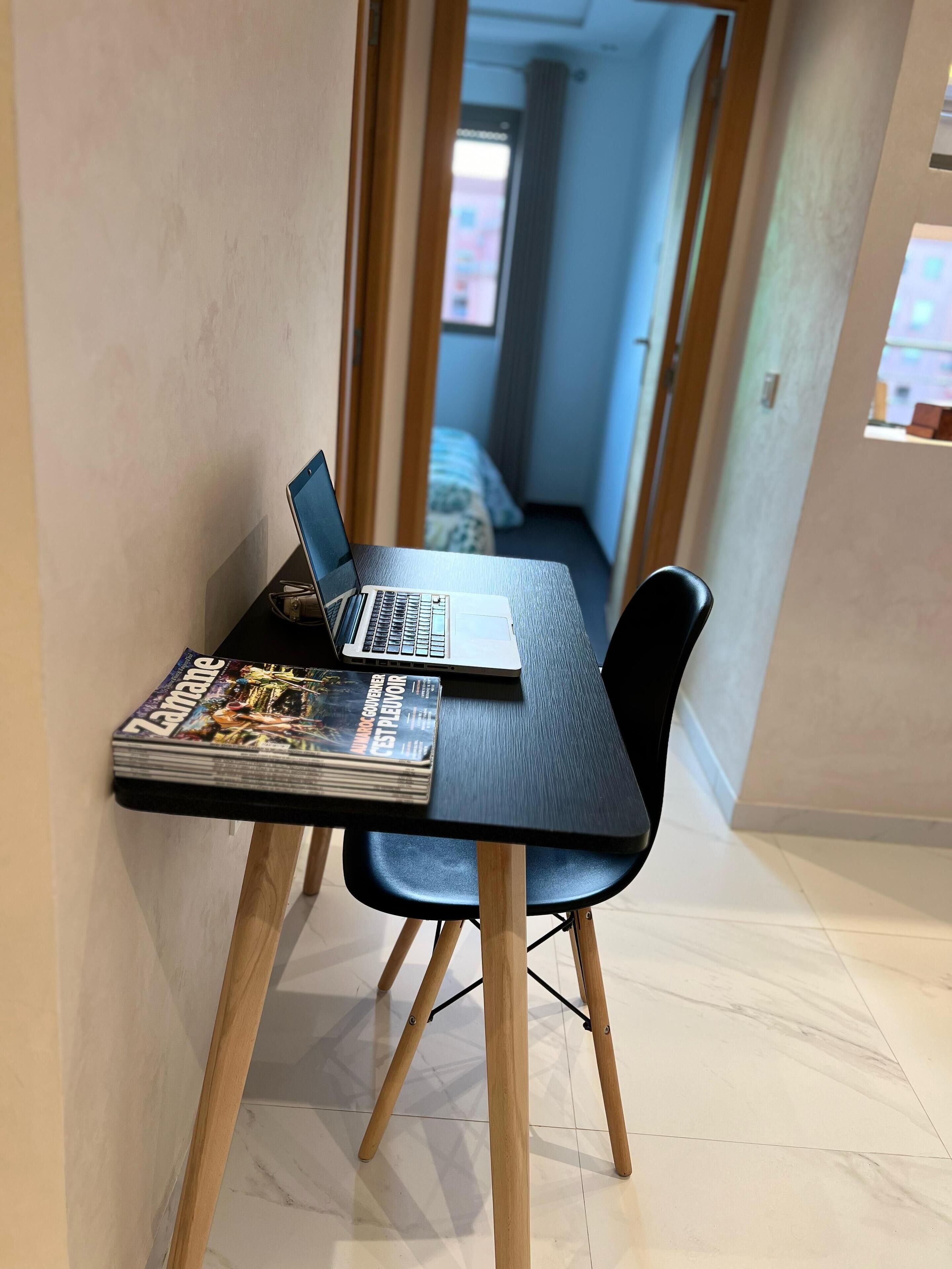 A small workspace with a laptop and a magazine on a table, a chair, and a view into a bedroom with a bed and curtains.