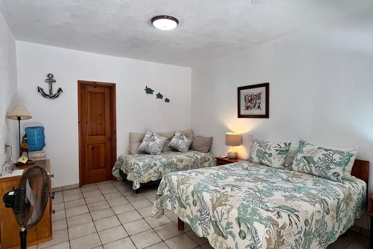 A bedroom with two beds, a wooden door, a blue water dispenser, a fan, and a tiled floor.