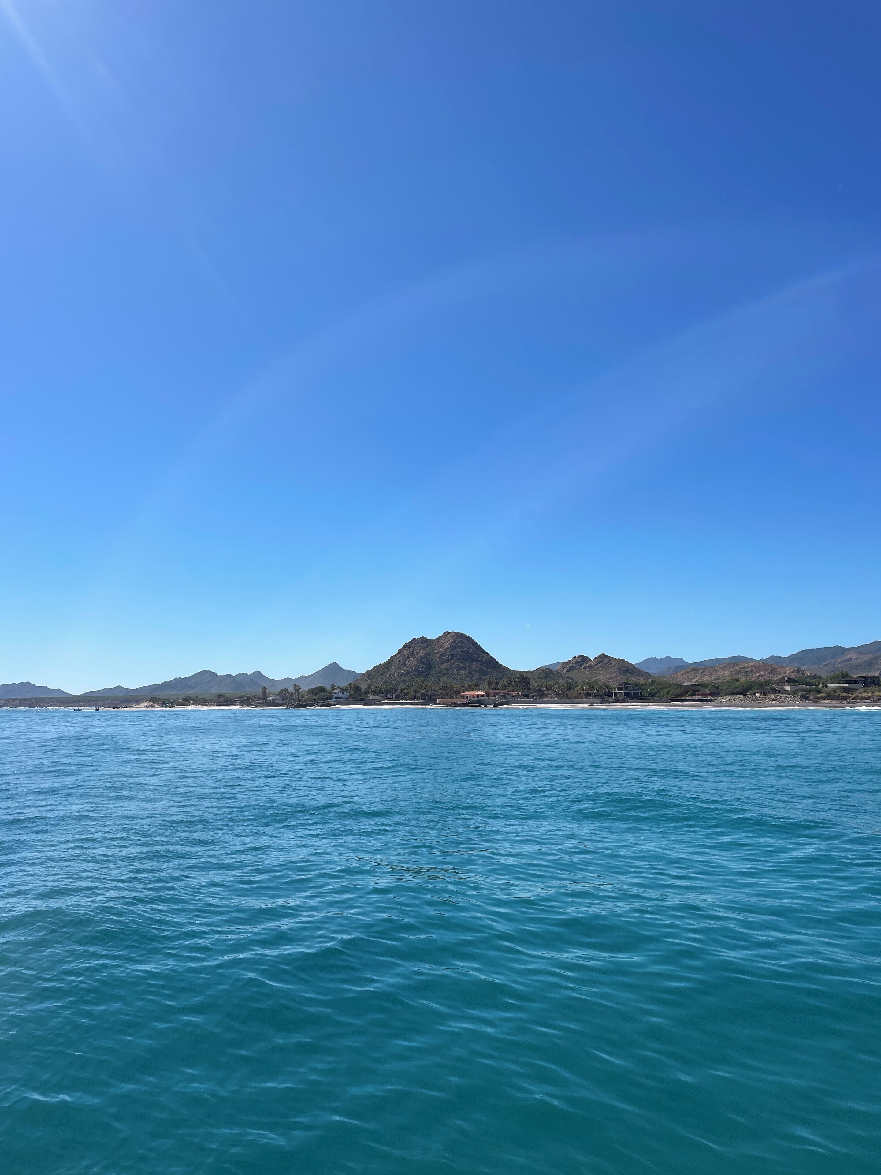 Eine Küstenlandschaft mit klarem, blauem Wasser, einem Gebirgszug und einem Strand mit Gebäuden.