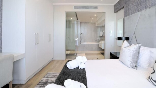 Una habitación de hotel moderna con una cama grande, una mesita de noche, un armario y un baño visible a través de una puerta de cristal.