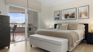 A bedroom with a bed, a bench, a nightstand, and a window with a view of buildings. - Apartment in Terrazas de Banús for 4 people | 13-4-1 (Puerto Banús - Marbella)